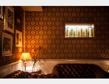 Wohnzimmerähnlicher Raum mit dunkelroter Tapete und goldenen Mustern, einer braunen Ledercouch und einer Tischlampe auf einem Beistelltisch mit einer Vase voller Trockenblumen. An der Wand hängen gerahmte Bilder, und ein beleuchtetes Regal zeigt Flaschen oder Behälter.