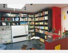 Moderne Bibliothek in einem Wohnraum mit schwarzen, deckenhohen Bücherregalen, die mit Büchern und Dekorationsobjekten gefüllt sind. Im Vordergrund befinden sich eine schwarze Liege, ein kleiner Tisch mit einem aufgeschlagenen Buch und eine rote Theke mit dekorativen Kerzen und Gegenständen.