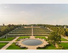 Weitläufige Gartenanlage mit terrassierten Weinreben, zentralem Weg und einem runden Wasserbecken im Vordergrund. Im Hintergrund befindet sich das Schloss Sanssouci mit einer grünen Kuppel, umgeben von Bäumen; links ist eine historische Windmühle zu sehen.