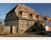 Backsteinhaus mit steilem Satteldach und mehreren Gaubenfenstern, teils mit roten Holzverkleidungen. Das Haus steht an einer gepflasterten Straße, umgeben von einem kleinen Zaun und Bäumen im kahlen Winterzustand.