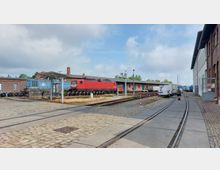 Bahnbetriebshof mit Gleisanlagen und einem roten Güterzug neben einem blauen Waggon. Umliegende Gebäude aus rotem Backstein und Weiß; im Vordergrund Kopfsteinpflaster sowie parkende Fahrzeuge.