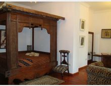 Schlafzimmer mit einem hölzernen Himmelbett im traditionellen Design, verziert mit Schnitzereien. Der Raum hat Holzboden, gemusterte Möbel, darunter Stühle und eine Couch mit Leopardenmuster, sowie minimalistische Wanddekorationen.