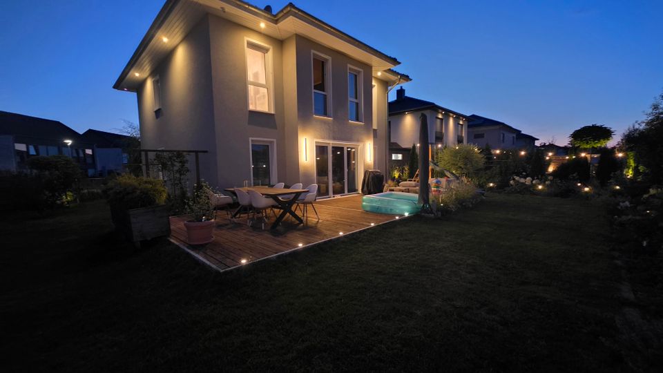 Beleuchteter Garten hinter einem modernen zweigeschossigen Einfamilienhaus in der Dämmerung, mit Holzterrasse, Esstisch und Stühlen sowie Einbauspots im Deck und Fassadenleuchten. Rasenfläche mit Sträuchern und Gartenbeleuchtung, im Hintergrund weitere Wohnhäuser.