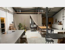 Offener, loftartiger Innenraum mit Betonboden und sichtbaren Deckenleitungen, ausgestattet mit langen Arbeits- und Esstischen, Hockern und einer Küchenzeile an der rechten Wand. In der Mitte trennt ein beiger Vorhangbereich mit Pflanzen und Rollwagen den Raum; links steht eine große Kücheninsel mit Arbeitsfläche.