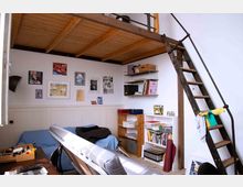 Kleines Arbeitszimmer mit einer Holzempore und einer Metalltreppe. An der weißen Wand hängen gerahmte Bilder, während Regale mit Büchern, Ordnern und Dekorationsgegenständen gefüllt sind; ein Schreibtisch, eine Couch und weitere Möbel ergänzen den Raum.