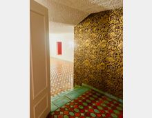 Ein Innenraum mit gemustertem Boden aus runden, bunten Fliesen in Grün, Rot und Orange. Eine Wand ist mit gold-orientalisch floralem Muster verziert, während die andere weiß verputzt ist und eine rote, rechteckige Nische enthält.