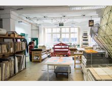 Große Werkstatt mit hohen Decken, in der eine Person an einem Tisch arbeitet. Der Raum ist mit Regalen voller Materialien, Druckgeräten, großen Tischen und Racks für Kunstdrucke ausgestattet; im Hintergrund befinden sich große Fenster, die Tageslicht hineinlassen.