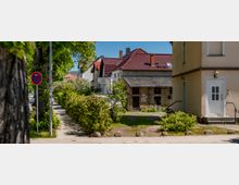 Wohnstraße mit schmalem Gehweg entlang großer Straßenbäume und Hecken, daneben ein kleines Grundstück mit altem Backstein-Nebengebäude. Im Hintergrund stehen Wohnhäuser mit roten Ziegeldächern; vorne rechts ist der Eingang eines beigefarbenen Hauses zu sehen.