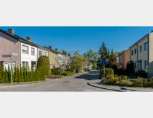 Eine ruhige Wohnstraße mit Reihenhäusern in unterschiedlichen Farben, gepflegten Vorgärten und blühenden Bäumen. Am Straßenrand steht ein Verkehrsschild und im Hintergrund sind Bäume und Vegetation sichtbar.