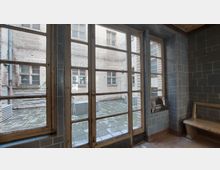 Blick durch eine verglaste Metalltür in einen Innenhof mit gepflastertem Boden und hellen Backsteinfassaden. Der Raum innen ist mit blauen Fliesen verkleidet und enthält eine Sitzbank sowie ein kleines Fenster.