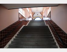 Treppenaufgang in einem historischen Gebäude mit braunen Wandfliesen und cremefarbenen Wänden. Am oberen Ende der Treppe befinden sich ein großes Rundbogenfenster und eine dekorative Lampe.