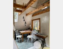 Gemütlicher Wohnraum mit Holzbalkendecke, einem kleinen Esstisch und zwei Stühlen, umgeben von hellem Holzvertäfelung. Ein Fenster mit Holzrahmen lässt Tageslicht herein, während ein dekorativer Teppich den Boden bedeckt; daneben steht ein Vintage-Nähmaschinentisch.