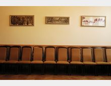 Reihe von braunen Holzstühlen mit gepolsterten Sitzen an einer beigefarbenen Wand, die mit drei gerahmten Bildern dekoriert ist; Holzboden mit Fischgrätenmuster darunter.