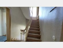 Treppenhaus in einem älteren Gebäude mit hölzernem Geländer, leicht abgenutzten Stufen und einer Wand mit weißer Strukturfarbe; ein Fenster am oberen Ende sorgt für Tageslicht. Links ist der Eingang zu einem weiteren Raum sichtbar.