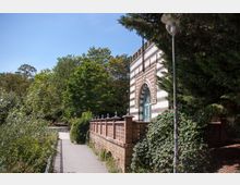 Backsteinbau mit gemustertem Rundbogen vor einem Weg, der durch eine grüne Parklandschaft mit Bäumen und Sträuchern führt. Rechts steht eine Laterne, während die Fassade durch ihre cremefarbenen und roten Streifen sowie dekorative Elemente hervorsticht.