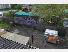 Auf dem Bild ist ein ländliches Gebiet mit einem kleinen eingezäunten Hof zu sehen. Im Vordergrund stehen ein grauer Hühnerstall mit orangefarbenem Wabenmuster und ein Gebäude aus Holz mit der Aufschrift „Don’t tick up!“. Im Hintergrund befinden sich ein Traktor, Bäume und weitere einfache Schuppen.