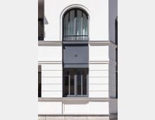 Fassade eines modernen Gebäudes mit heller Putzoberfläche, hervorgehoben durch zwei Fenster: ein oben gelegenes, rundbogiges Fenster und ein rechteckiges Fenster darunter. Beide Fenster haben dunkle Rahmen und Balkongitter.