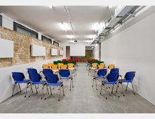 Ein moderner Seminarraum mit freiliegender Backsteinwand und Betonboden, bestückt mit bunten Stühlen (blau, gelb, rot, grün), die auf einen Projektor und eine Leinwand ausgerichtet sind. An der Decke verlaufen sichtbare Leitungen und Lampen, die den Raum gleichmäßig beleuchten.