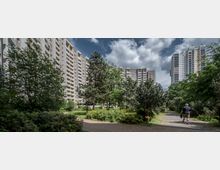 Wohnkomplex mit mehreren hohen, hellen Plattenbauten, umgeben von grünen Bäumen, Sträuchern und einer parkartigen Anlage mit Gehwegen; eine Person spaziert auf einem der Wege.