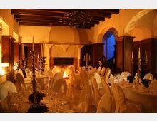 Eleganter Speisesaal mit festlich gedeckten runden Tischen und weißen Stuhlhussen, beleuchtet von warmem Kerzenlicht und einem Kamin im Hintergrund. Der Raum hat eine Holzdecke mit Balken, holzverkleidete Wände und einen hellen Steinbogen, der zu einem blauen Bereich führt.