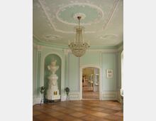 Eleganter, klassischer Innenraum mit mintgrünen Wänden, verziert mit weißen Stuckelementen und einem Kristallleuchter an der reich dekorierten Decke. Der Raum hat einen Parkettboden, einen großen Kachelofen an der Wand und eine geöffnete Tür, die zu einem angrenzenden Bereich mit Ausstellungsstücken führt.