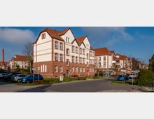 Ein Wohngebiet mit großen, mehrstöckigen Gebäuden aus rotem und hellem Backstein im Stil der klassischen Architektur. Im Vordergrund befinden sich geparkte Autos, eine Straße und mehrere Verkehrs- sowie Straßenschilder; im Hintergrund ist ein Schornstein sichtbar.