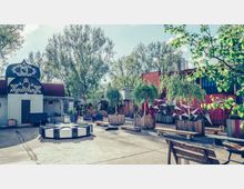 Ein urbaner Außenbereich mit bunt bemalten Containern, kleinen Holzbauten und zahlreichen bepflanzten Holzbehältern. Im Vordergrund stehen Sitzbänke und ein achteckiger Tisch, umgeben von Bäumen und grüner Vegetation.