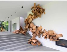 Moderne Innenraumaufnahme mit einer großen, organisch geformten Holzskulptur, die eine weiße Wand sowie die angrenzende Decke und den Boden entlang kriecht. Im Hintergrund befinden sich Glasfenster, die eine grüne Landschaft zeigen, sowie eine Treppe.