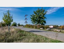 Asphaltierter Weg in einer ländlichen Umgebung mit einer Wiese im Vordergrund, einem einzelnen Baum und einer Hecke rechts. Links steht eine hölzerne maritime Markierung, und im Hintergrund sind Schilder und Bäume sichtbar unter einem klaren blauen Himmel.