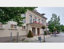 Außenansicht eines historischen Gebäudes mit der Beschriftung „Bibliothek“ über dem Eingang. Das Gebäude hat helle Fassade, große Fenster, ein Vordach mit einer Uhr und ist von Blumenarrangements sowie Bäumen auf einem gepflasterten Platz umgeben.