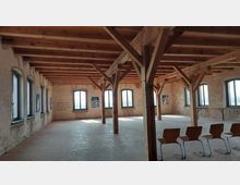 Ein heller Raum mit rustikaler Inneneinrichtung, gekennzeichnet durch sichtbare Holzbalken an der Decke sowie Backsteinwände mit mehreren großen, bogenförmigen Fenstern. Es stehen einige Stühle in einer Reihe, und an den Wänden hängen mehrere gerahmte Bilder.