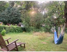 Grüner Garten mit Rasenfläche, dichtem Busch- und Baumbestand; im Vordergrund stehen Holzstühle, rechts hängt eine Hängematte an einem Baum. Im Hintergrund sind niedrige Gebäude teilweise hinter Hecken und Bäumen sichtbar, mit Sonnenlicht, das durch das Laub fällt.