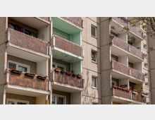 Mehrstöckiges Wohnhaus im Stil eines Plattenbaus mit Balkonen, die teils braune Fliesenverkleidungen aufweisen. Einige Balkone sind mit Blumenkästen dekoriert, während die Fassade sichtbare Abnutzungsspuren zeigt.