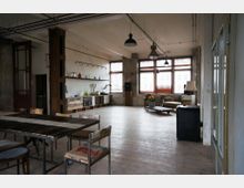 Ein geräumiger, industriell gestalteter Raum mit Holzboden, großen Fenstern mit Jalousien und sichtbaren Deckenrohren. Links steht ein langer Holztisch mit Stühlen, im Hintergrund eine offene Küche mit rustikalen Holzschränken und Regalen; rechts eine Sitzecke mit Sofas, Sesseln und einem niedrigen Couchtisch.