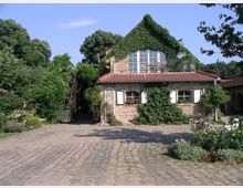 Backsteinbau mit roten Ziegeldächern, teils von Efeu bewachsen, umgeben von einem gepflasterten Hof und üppiger Vegetation. Das Gebäude verfügt über weiße Fenster mit grünen Fensterläden sowie einen Balkon im Obergeschoss.