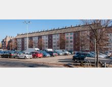 Mehrstöckiges Wohngebäude mit Satteldach im Hintergrund, gebaut aus roten und beigen Fassadenteilen. Im Vordergrund befindet sich ein großer Parkplatz mit geparkten Autos, beleuchtet von einer einzelnen Straßenlaterne, sowie einigen kahlen Bäumen.