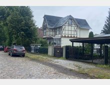 Fachwerkhaus mit dunkelgrauem Schieferdach in einer ruhigen Wohnstraße, flankiert von Bäumen und geparkten Autos auf einem Kopfsteinpflasterweg. Ein schmiedeeiserner Zaun und ein Carport befinden sich im Vordergrund.