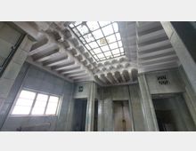 Innenansicht eines Raumes mit geometrisch gestaltetem Kassettendeckenmuster und verglaster Oberlichte, die durch Schmutz teilweise verdeckt ist. Wände aus hellen, glasierten Kacheln mit schmalen Lüftungsöffnungen und einer Fensterfront auf der linken Seite.