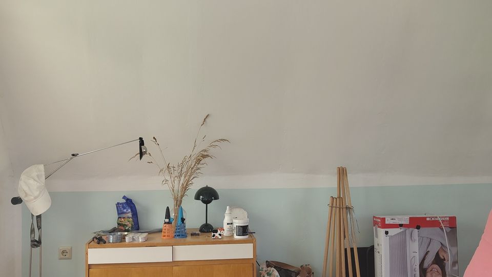 Innenraum eines Dachzimmers mit schräger weißer Decke und sichtbaren Deckenbalken, Holzfußboden und zweifarbig gestrichener Wand. Vor der Wand steht ein niedriges Holzsideboard mit Gegenständen und Lampe; rechts ist ein Bett mit blauem Bezug sowie mehrere abgestellte Taschen, Kartons und Stiefel zu sehen.