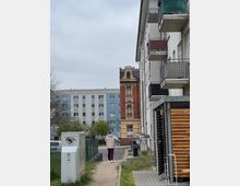 Ein Wohngebiet mit mehrstöckigen Gebäuden: rechts ein modernes Wohnhaus mit Balkonen, in der Mitte ein historisches, rot-gelbes Gebäude mit verzierten Elementen, und im Hintergrund ein graues Plattenbaugebäude. Im Vordergrund führt ein Gehweg durch eine grüne Fläche, daneben ein Stromkasten und eine Person, die weggeht.
