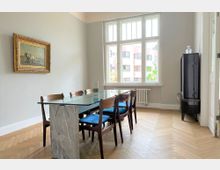 Esszimmer mit großem Glastisch auf einem Steinsockel und sechs Holzstühlen mit blauen Sitzkissen, Parkettboden in Fischgrätmuster, große Fenster mit Blick auf ein gegenüberliegendes Gebäude und eine verzierte Rahmung an der Wand mit einem Gemälde.