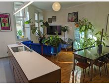 Wohnzimmer mit offener Küche und Holzboden, dekoriert mit zahlreichen Zimmerpflanzen. Im Raum befinden sich ein blauer Samtsofa, eine lange Kücheninsel, ein Esstisch mit Metalloberfläche und Stühlen sowie Wände mit Kunstwerken und großen Fenstern, die viel Tageslicht hereinlassen.