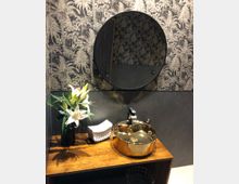 Ein stilvoll gestaltetes Badezimmer mit einer goldenen, runden Waschschale auf einer hölzernen Ablagefläche, einem schwarzen Wasserhahn und einer Vase mit weißen Lilien. Der Hintergrund zeigt eine dunkle Tapete mit Palmenmotiven.