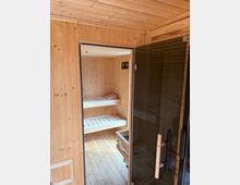 Wood-paneled sauna room with a wooden plank floor and ceiling, viewed through a glass door with a vertical handle. Inside are two-tier wooden bench seats along the back wall and a small heater unit near the entrance.