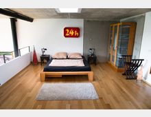 Schlafzimmer mit modernem Design, ausgestattet mit einem Holzbett, zwei Nachttischen, einer Glasvitrine und Holzboden. Eine Betonwand und eine weiße Wand mit einem roten Schild "24h" ergänzen das minimalistische Ambiente.