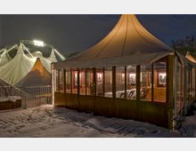 Ein beleuchtetes Festzelt mit gläsernen Wänden und Holzrahmen steht in einer verschneiten Umgebung. Im Hintergrund sind größere weiße Zirkuszelte mit Lichterketten und einer leuchtenden Schrift sichtbar.