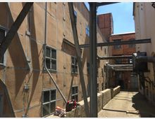 Industriekomplex mit zwei Gebäuden, eines mit hellen verputzten Wänden und das andere aus rotem Backstein. Sichtbare Metallträger, kleine Fenster und Rohrleitungen prägen die Szene, während Schienen in den gepflasterten Boden eingelassen sind.