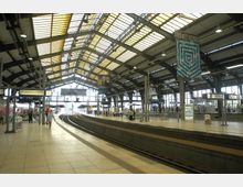 Bahnhofshalle mit gewölbtem Glasdach, darunter mehrere Gleise und Bahnsteige. Menschen stehen oder gehen auf den Bahnsteigen, Werbetafeln und Beleuchtung hängen an der Stahlkonstruktion.