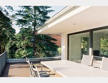 Moderne Terrasse mit Holzboden und weißem Tisch, umgeben von Glasgeländern. Im Hintergrund sind Bäume und ein Haus mit grünem Anstrich und roten Dachziegeln zu sehen.