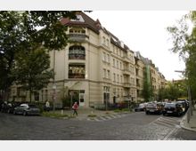 Mehrstöckiges Wohngebäude mit heller Fassade und Erkern an einer Straßenecke, umgeben von Bäumen. Geparkte Autos stehen entlang der gepflasterten Straße, während einige Personen auf dem Gehweg zu sehen sind.